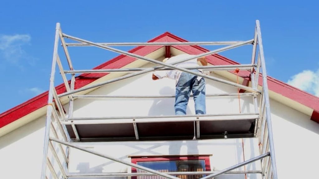 Een vakman legt vanop een stelling de laatste hand aan het verven van de gevelpleister. De rode dakgoot en het rode raam is perfect afgestemd op de witte crepi gevel.