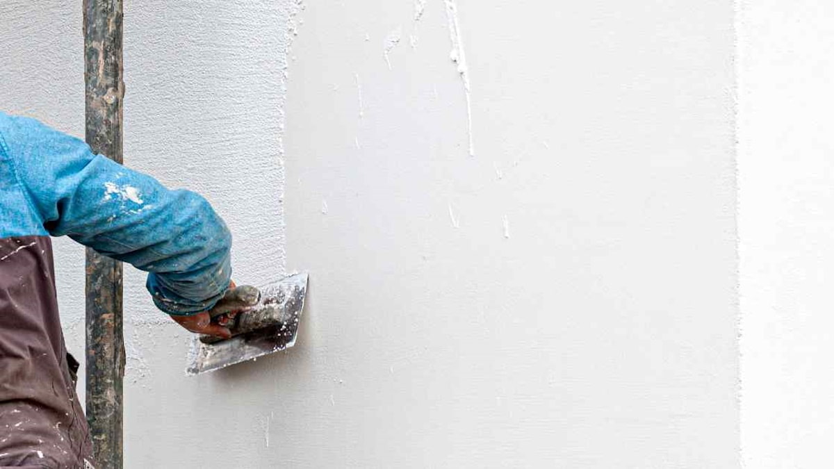 Een stukadoor brengt gevelpleister met een structuur aan op een gladde gevel. De man draagt beschermende kleding en gebruikt een spaan.