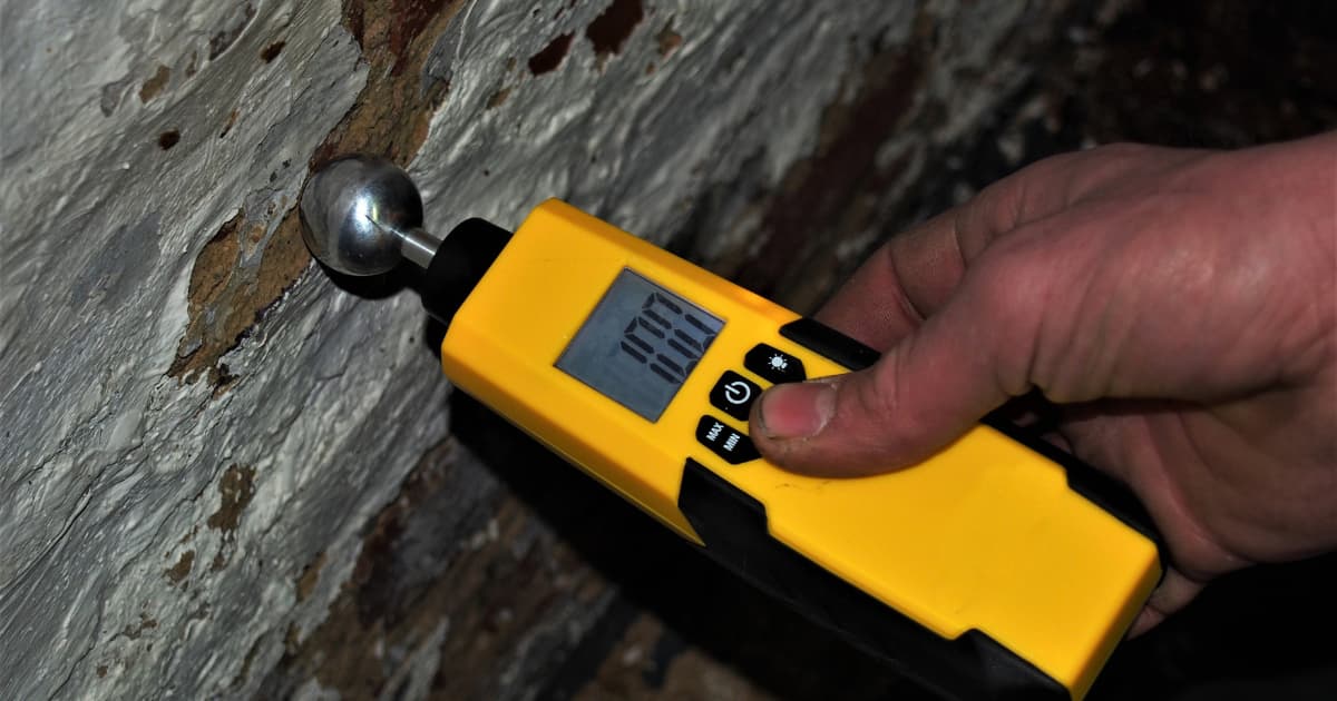 Een hand drukt de metalen bolsensor van een gele digitale vochtmeter tegen een oude muur met afbladderende witte verf.