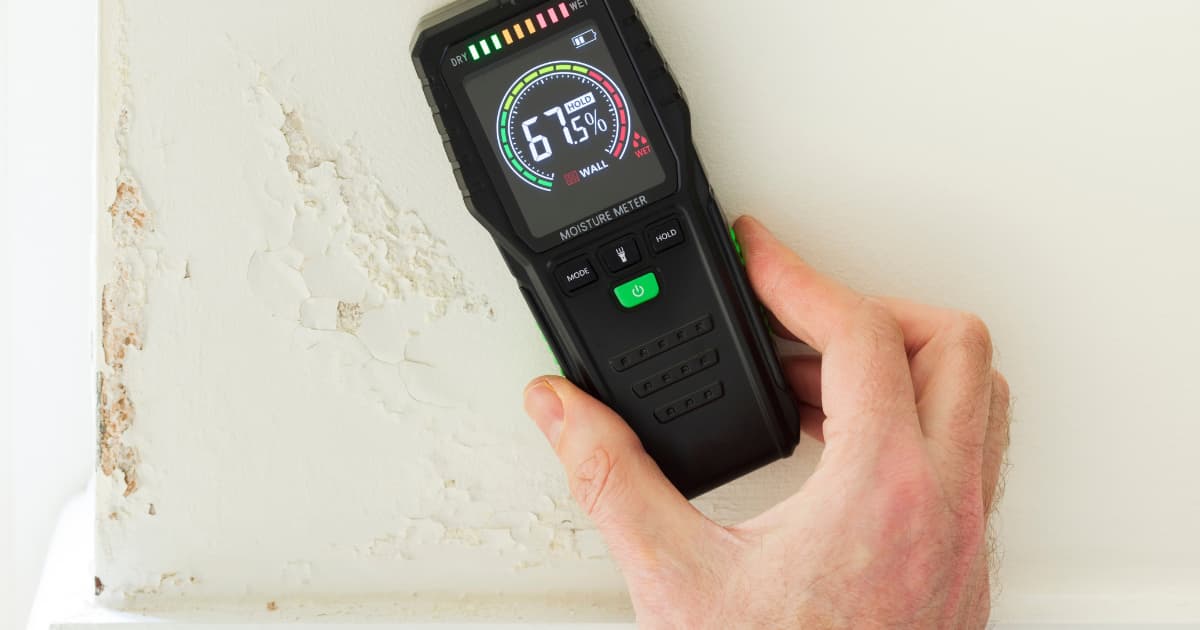 Een hand houdt een zwarte pinloze vochtmeter met kleurenschaal van droog naar nat tegen een witte muur met gebarsten en afbladderende verf.