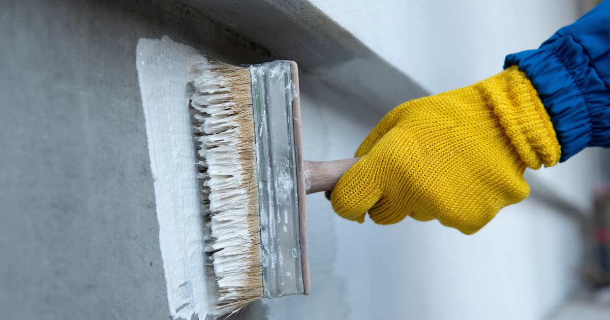 Een hand in een gele werkhandschoen brengt met een brede kwast een witte waterdichte coating aan op een grijze betonnen muur.