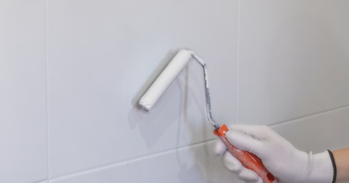 Een hand met een witte werkhandschoen rolt met een kleine verfroller met oranje handvat witte verf over grote wandtegels.