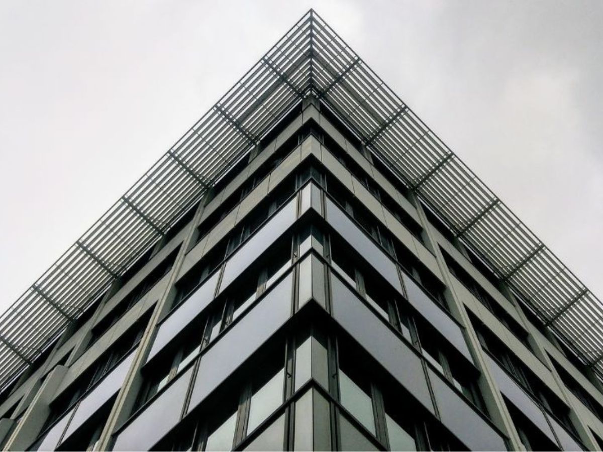 Hoek van een modern gebouw met glas- en metalen gevel en een metalen rooster bovenaan, gefotografeerd tegen een grijze lucht.