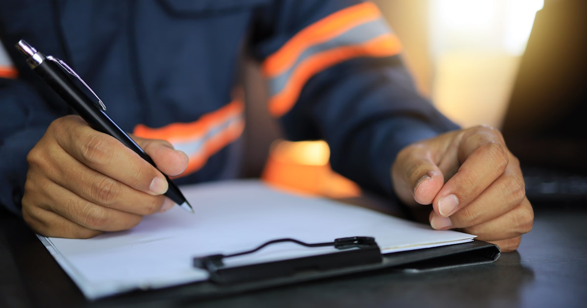 Een man schrijft op papier op een klembord. Hij draagt veiligheidskleding met oranje neon banden
