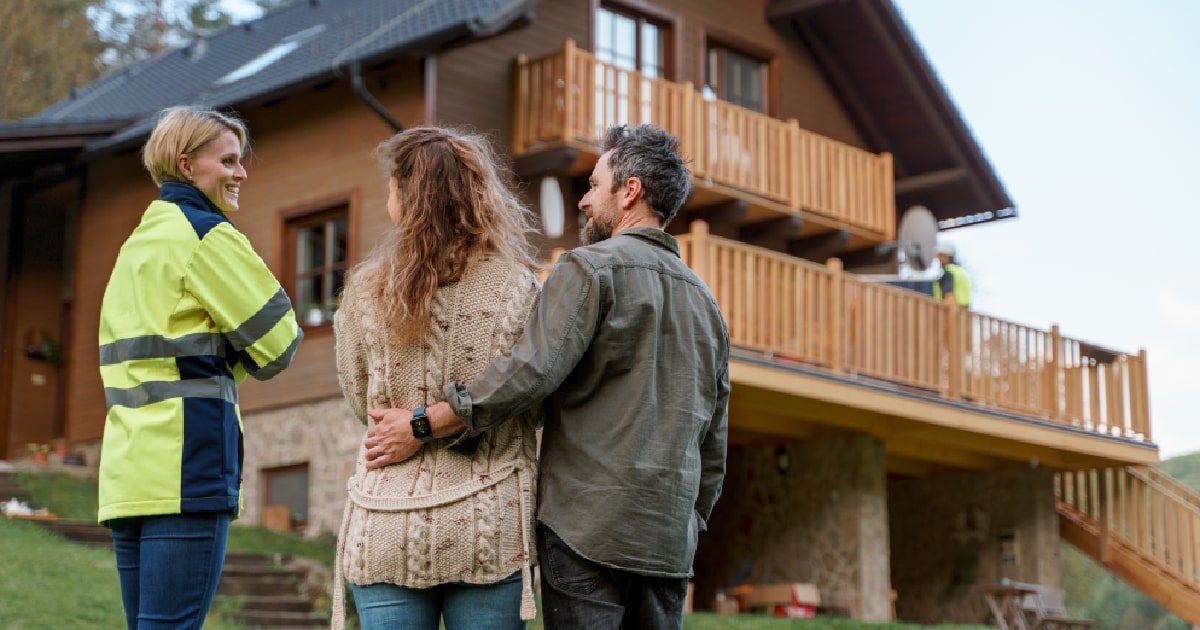 Een energiespecialist bespreekt de mogelijkheid tot een installatie met zonnepanelen en een thuisbatterij met een koppel, op de achtergrond staat een houten woning met verschillende verhoogde terassen.