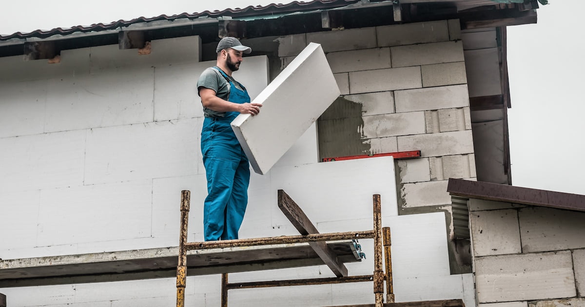 Een isolatiespecialist brengt isolatiemateriaal tegen de gevel van een woning aan. Hij staat op een steiger en draagt een overall