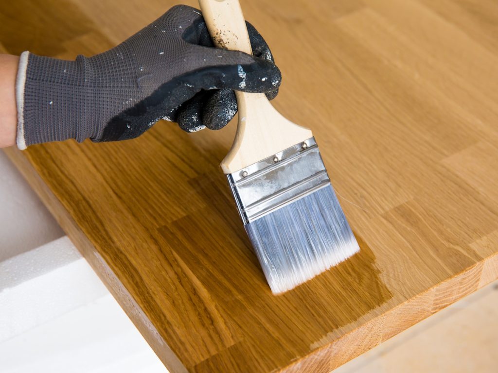 Een schilder lakt een aanrechtblad in een transparante kleur. De schilder draagt donkere handschoenen en gebruikt een platte blokkwast.