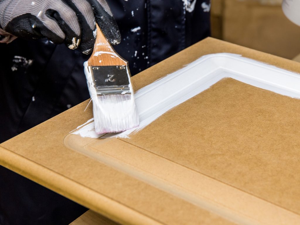 Een schilder verft een onafgewerkte deur van een keukenkastje wit. De schilder draagt een donkere broek en een handschoen en gebruikt een platte kwast.