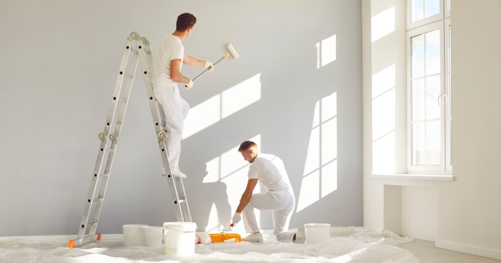 Twee schilders schilderen een muur met verfrollers. Ze dragen witte kleding. Een van de schilders staat op een trap, de ander knielt op de grond.