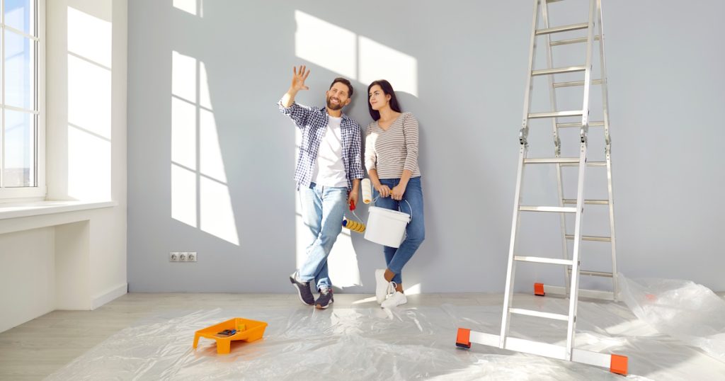 Een man en een vrouw staan in een ruimte die geschilderd moet worden. Ze hebben verfrollers en een verfemmer vast. De vloer is afgeplakt en er staat een ladder klaar.