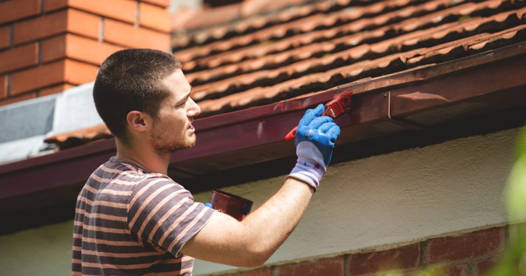 Een man verft de dakgoot van een huis. De man draagt een gestreept shirt en blauwe handschoenen. De dakpannen zijn oranje.