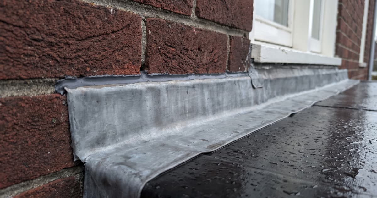 Een loodstrip sluit de overgang tussen een plat dak en een rode bakstenen gevel waterdicht af, met regendruppels zichtbaar op het natte dakoppervlak naast een wit kozijn.
