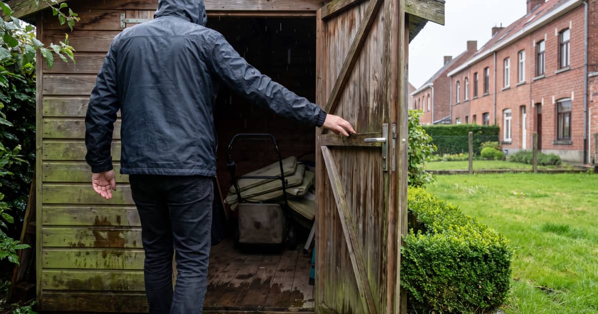 Een man in een donkere regenjas opent in de regen de deur van een houten tuinhuis met groene aanslag op de planken, waarbinnen opgestapelde tuinkussens op een donkere natte vloer liggen, terwijl een buxushaag en rode bakstenen rijhuizen op de achtergrond staan.