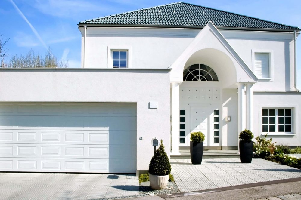 Een moderne, vrijstaande woning met een garage en een gevel van witte, minerale sierpleister. Er staan verschillende plantenpotten voor de ingang en op de oprit. 