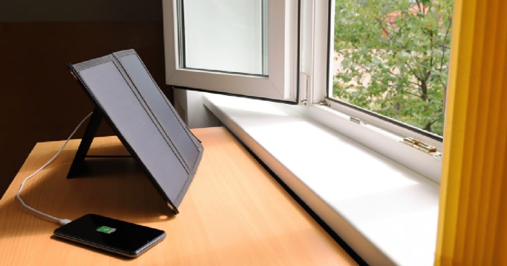 Een verplaatsbaar zonnepaneel vangt zon op door een geopend raam en laadt een smartphone op met groene stroom.