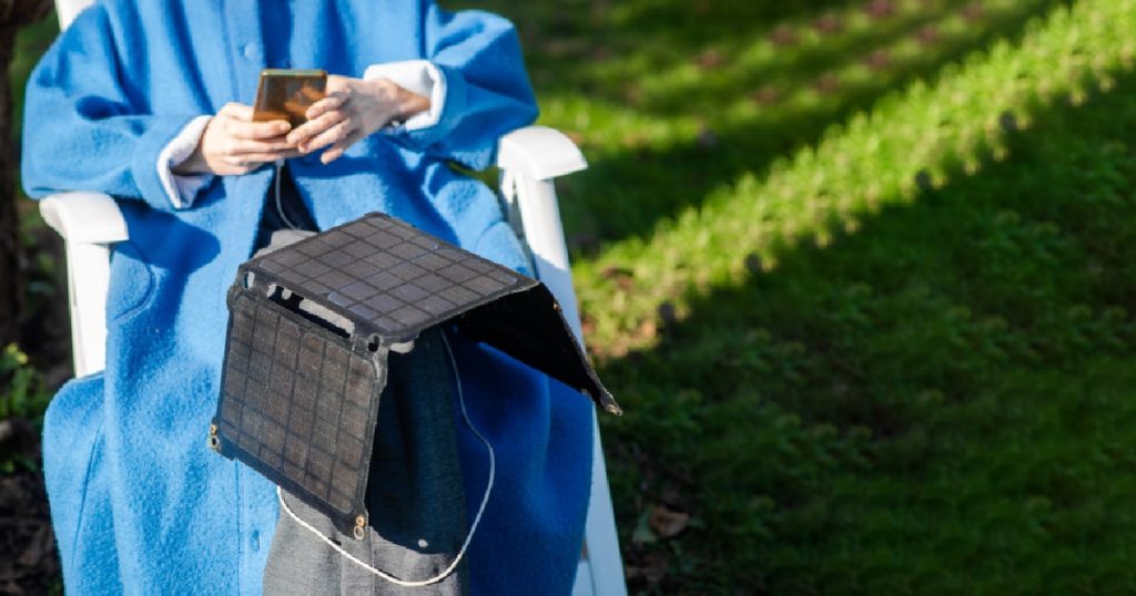 Een vrouw in een lange blauwe jas laadt haar smartphone op met groene energie dankzij haar mobiele thuisbatterij en mobiel zonnepaneel terwijl ze op een kampeerstoel zit.