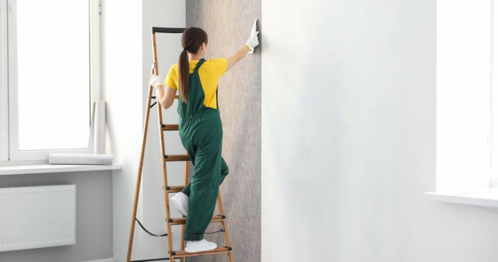 Een vrouwelijke behanger in groene overall en geel t-shirt gaat met een witte spatel over een net opgehangen strook behangpapier.
