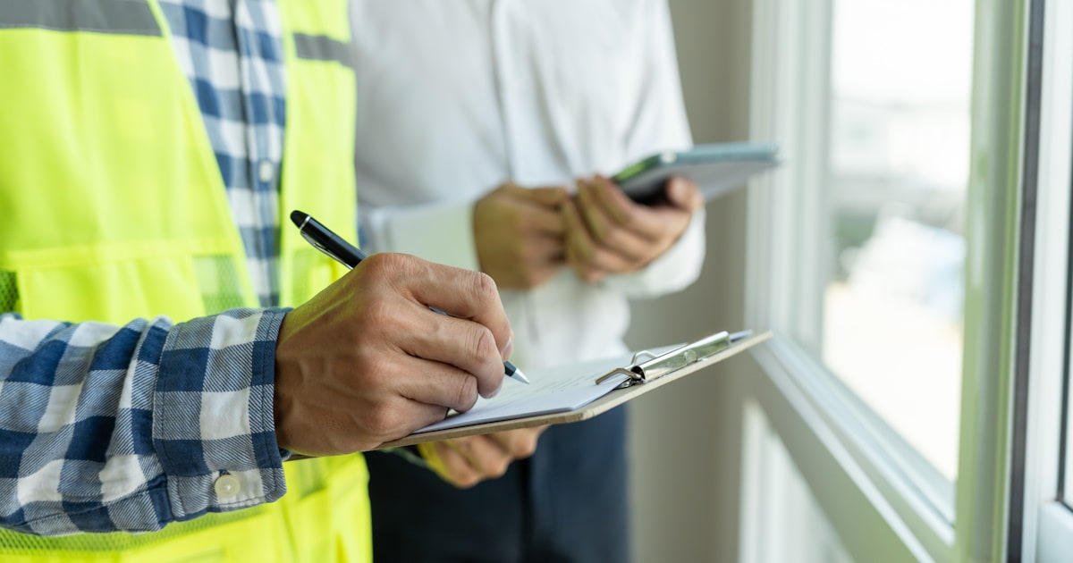 Een man in een veiligheidsvest schrijft op een klembord. Naast hem staat een man met een tablet in zijn handen