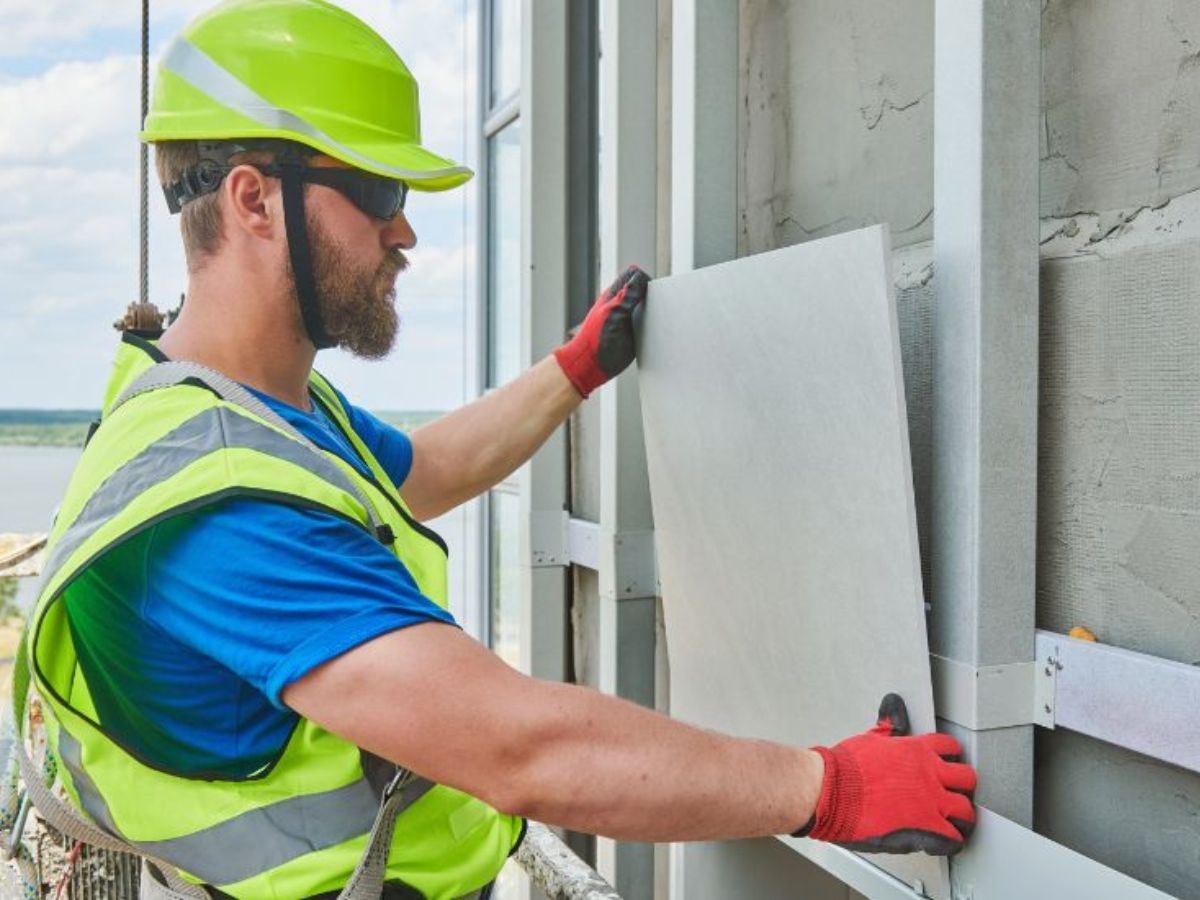 Bouwvakker met veiligheidshelm en handschoenen plaatst keramisch gevelpaneel op metalen onderconstructie.