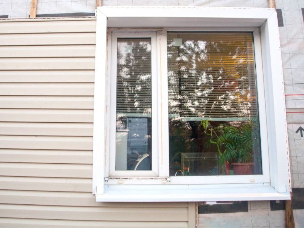 Detail van een woning tijdens de plaatsing van kunststof gevelbekleding in houtlook, rondom een wit kozijn met raam.