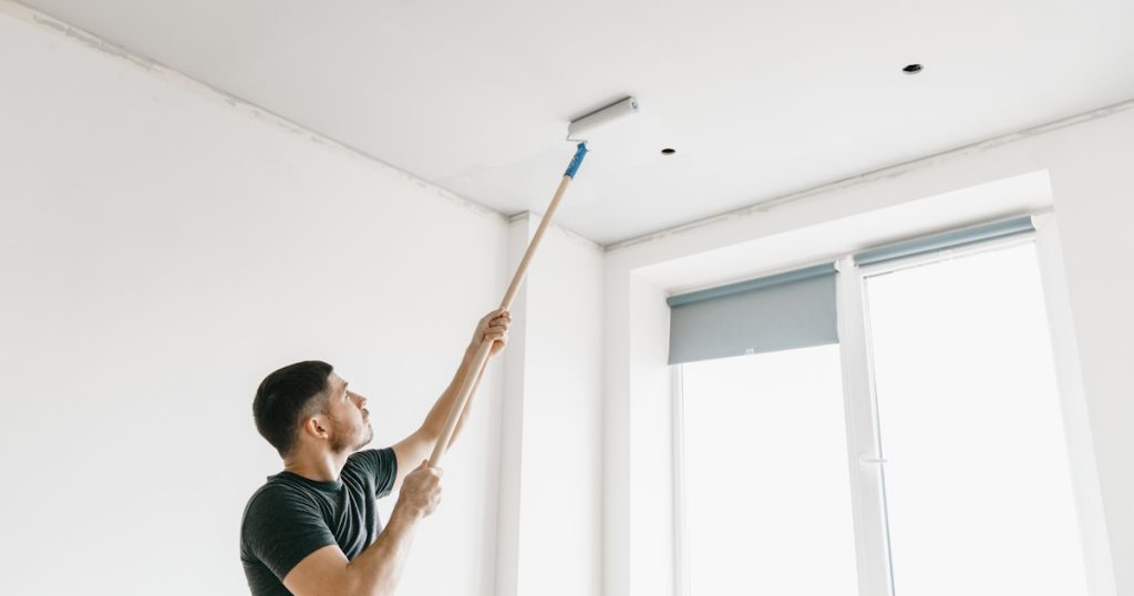 Een man wit het plafond met een telescopische verfroller. De man draagt een donker shirt. De ruimte is erg licht.
