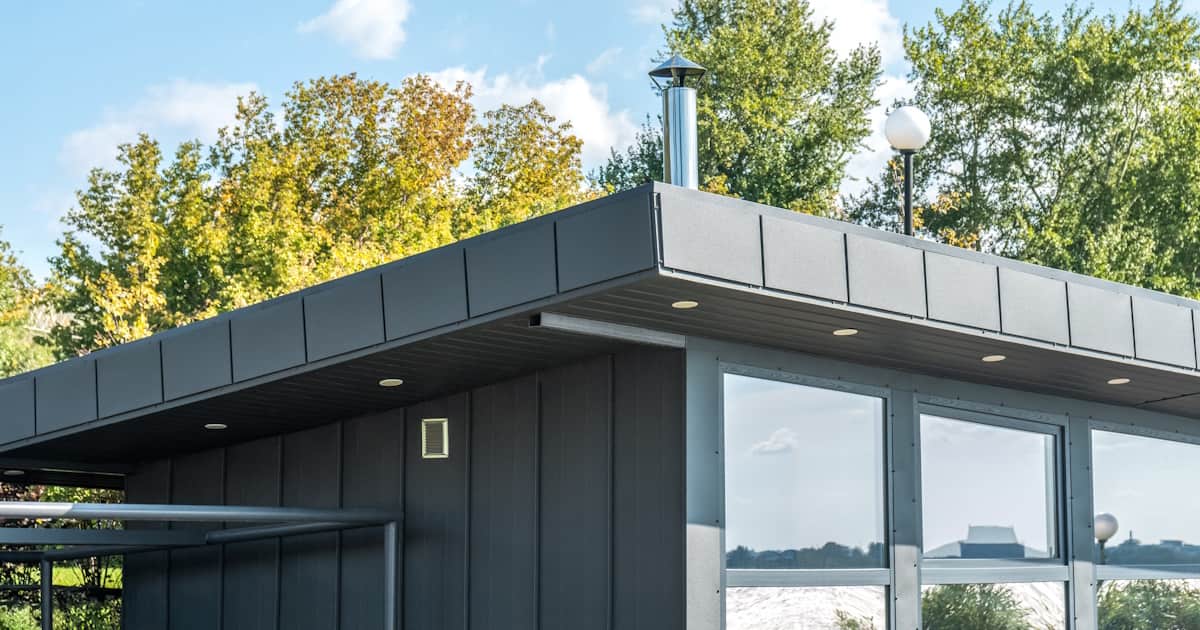 Het platte dak van een moderne woning. De woning is donker van kleur en is voorzien van grote ramen. Op de achtergrond bomen.