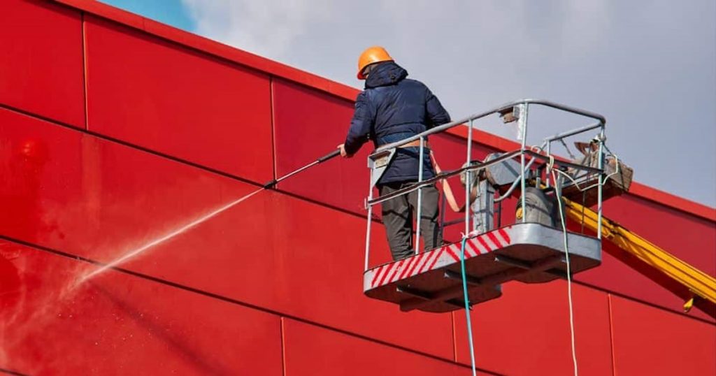 Voor het reinigen van een rode gevel wordt een hogedrukreiniger gebruikt. Omdat er op hoogte wordt gewerkt staat de vakman op een hoogwerker.