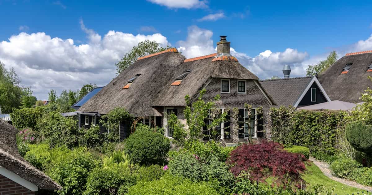 Een vrijstaande woning met een rieten kap. Op de voorgrond veel groen, op de achtergrond een bewolkte lucht.