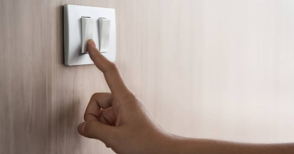 Een hand gebruikt de lichtschakelaar om de lampen van een woning uit te schakelen om zo het energieverbruik te beperken.