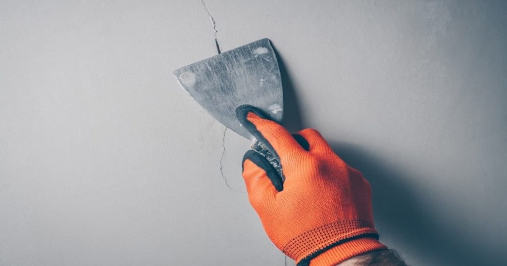 Een man herstelt een haarscheur in een muur met wat muurvuller en een plamuurmes. De man draagt een oranje handschoen.