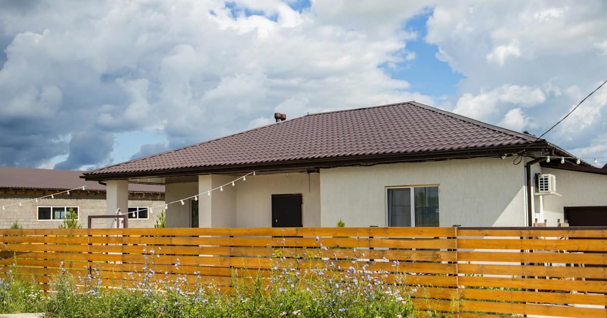 Een woning met een lichte gevel en een bruin schilddak. Voor de woning zijn een schutting en planten in bloei zichtbaar