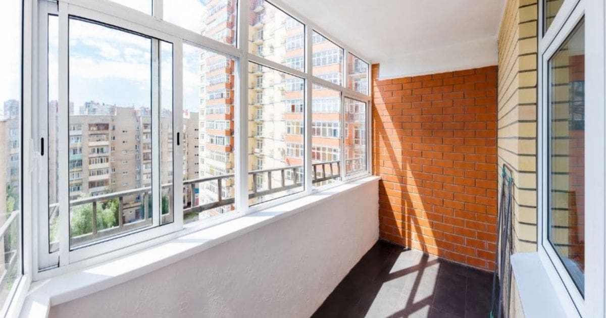 Een balkon met meerdere schuiframen met witte kozijnen. Door het raam kijken we uit op enkele flatgebouwen