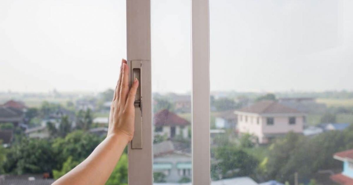 Een hand die een schuifraam dicht duwt, door het raam kijken we uit op een dorp met bomen en huizen