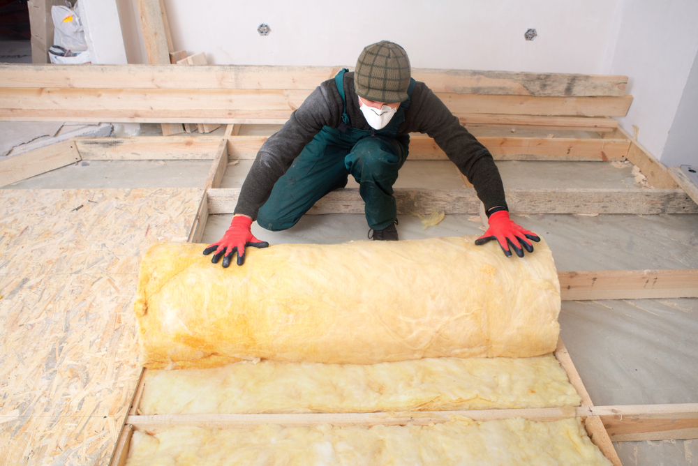 Een man legt vloerisolatie aan in een woning. De man rolt een stuk isolatiemateriaal uit en draagt rode handschoenen en een groene overall.