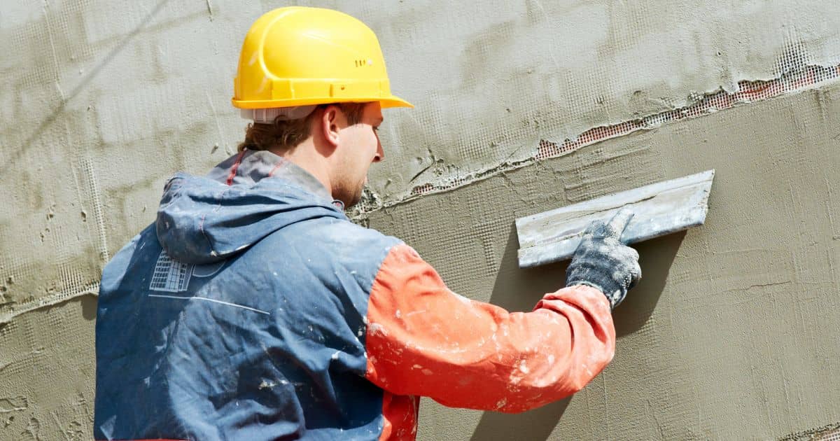 Een gevelspecialist met een gele veiligheidshelm brengt sierpleister aan over de wapeningslaag. Hij gebruikt er een spaan voor.
