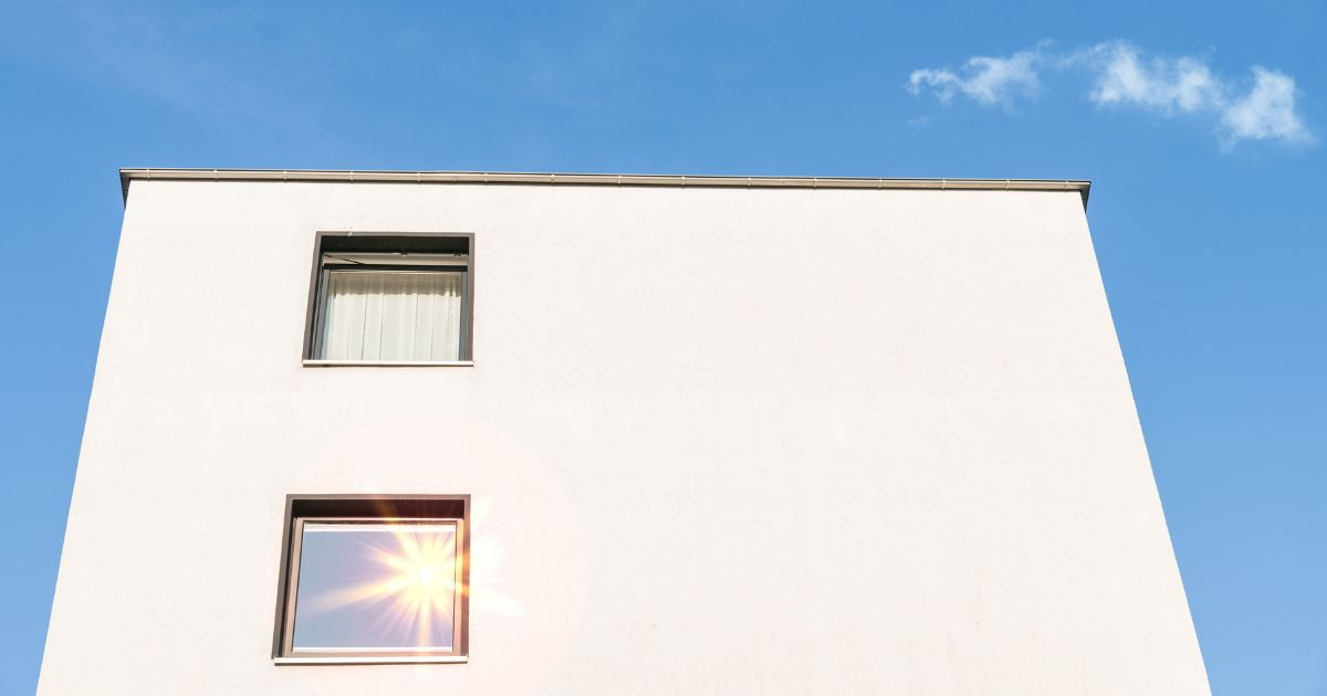 Een egale, witte sierpleister gevel steekt mooi af tegen een strakblauwe lucht. Links zijn twee raamkozijnen te zien.