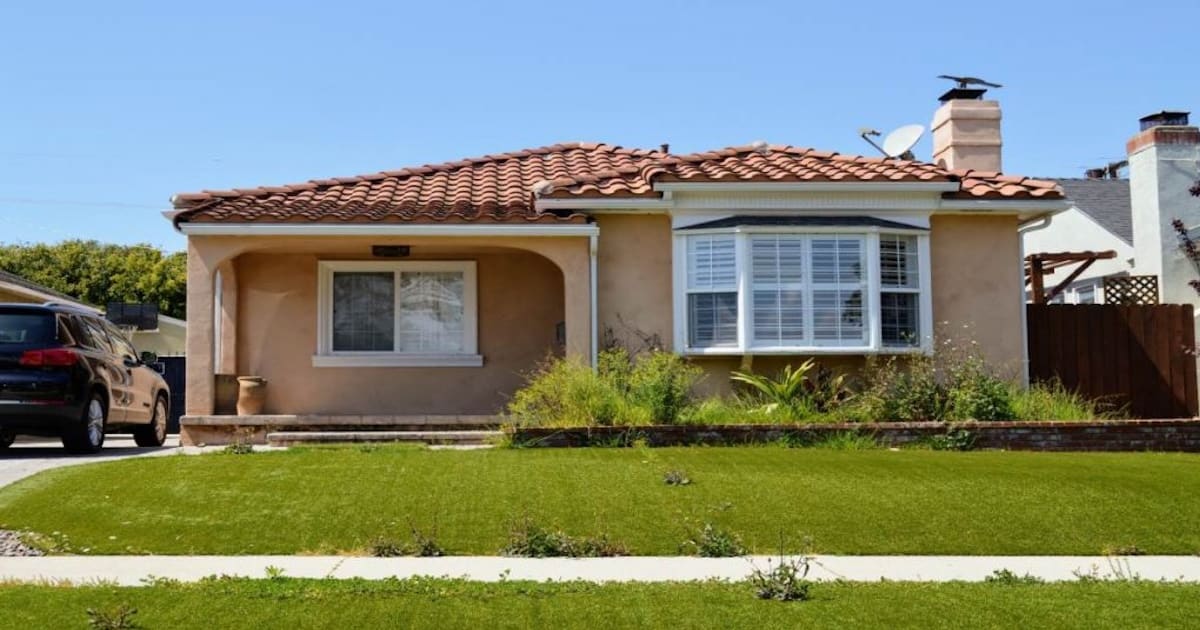 Een woning in een landelijke stijl met een gevel van een warme tint sierpleister. Op de voorgrond een gazon en aan de linkerkant van het huis staat een donkere auto geparkeerd. 