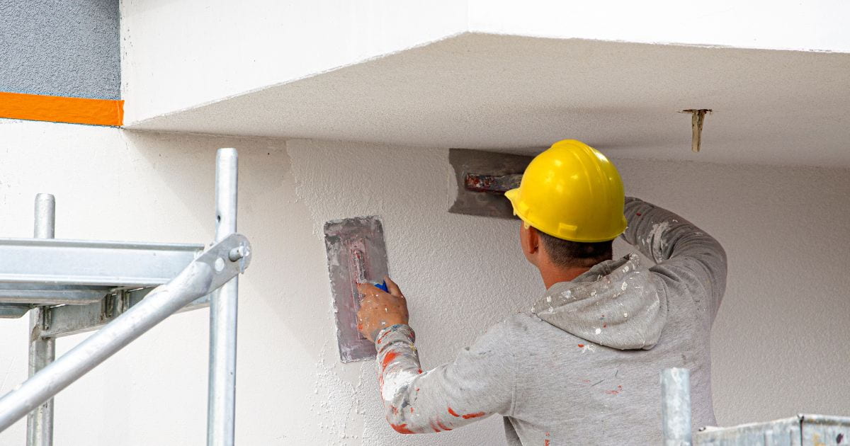 Een gevelspecialist met een gele helm brengt sierpleister met een grove korrel aan op een gevel.