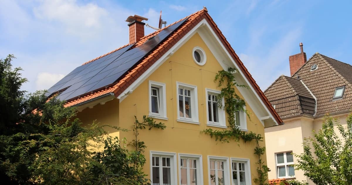 Een woning met een gele sierpleister gevel. Op de gevel zijn draden aangebracht om klimplanten tegenaan te laten klimmen. Op het dak liggen zonnepanelen.