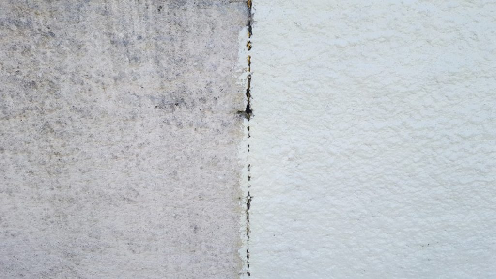 Een sierpleister gevel die voor de helft is geschilderd. Links is de gevel nog grijs, terwijl de muur rechts een witte laag verf heeft gekregen.