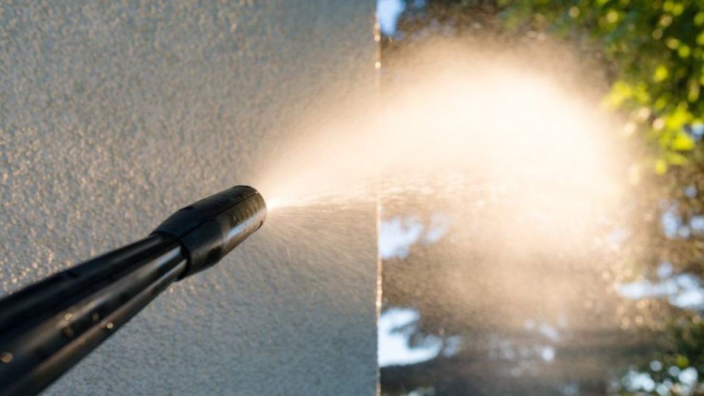 De sierpleister op de gevel wordt gereinigd met een hogedrukreiniger. Op de achtergrond is een boom te zien.
