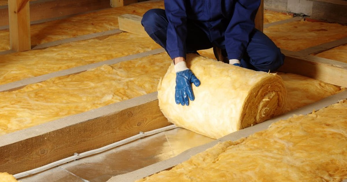 Een isolatiespecialist plaatst een rol minerale wol om een ruimte te isoleren. De specialist draagt een beschermende, donkere overall en handschoenen.