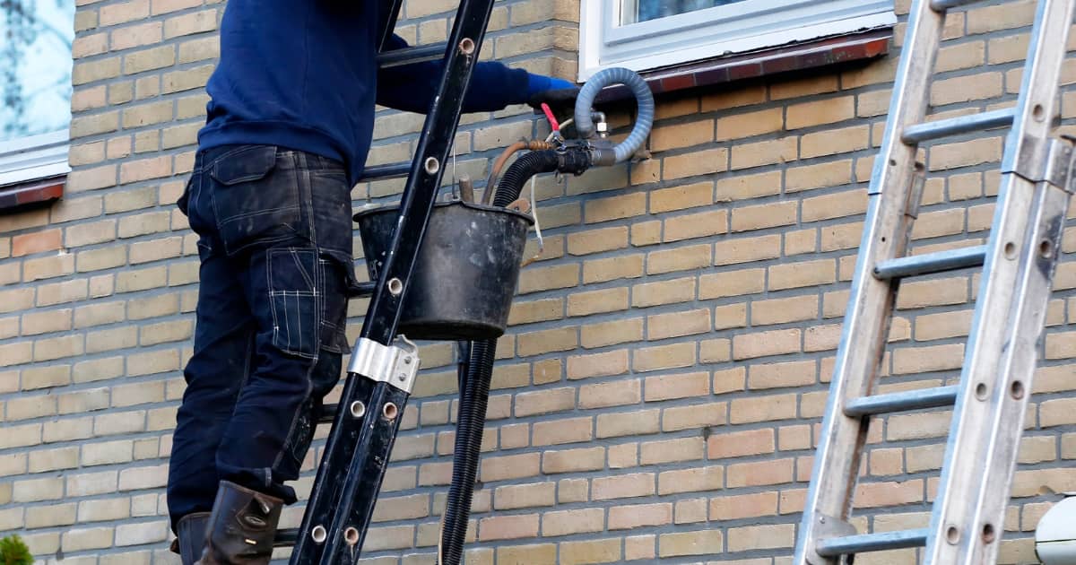 Een vochtspecialist staat op een ladder tegen een lichtgele bakstenen gevel en spuit isolatiemateriaal via een slang in een boorgat in de spouwmuur.