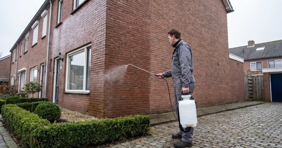 Een specialist in grijs werkpak sproeit met een drukspuit impregnatiemiddel op de bakstenen gevel van een Nederlands rijtjeshuis terwijl een fijne nevel zichtbaar de muur bedekt.