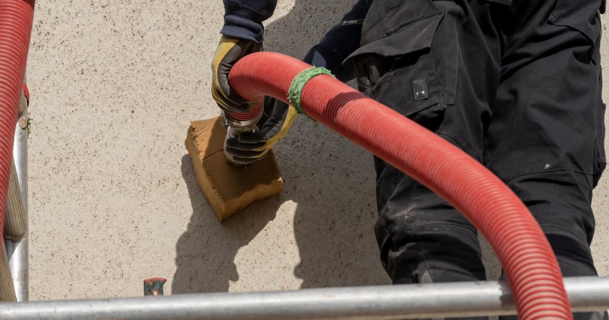 Een specialist blaast met een rode slang isolatiemateriaal in de spouwmuur. De expert draagt handschoenen en een werkbroek