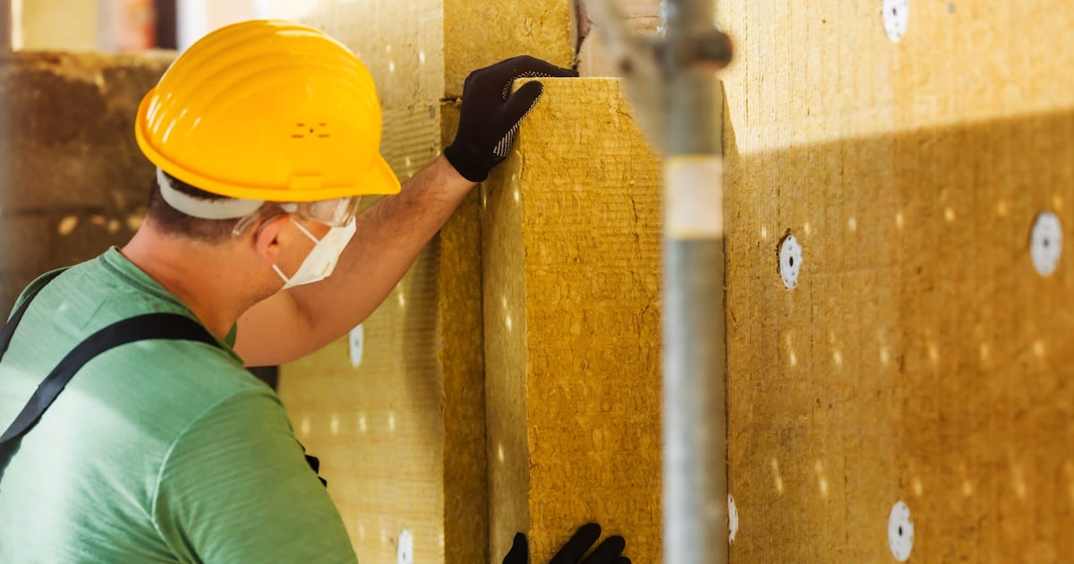 Een isolatiespecialist brengt steenwol isolatie aan. Hij draagt een veiligheidshelm, mondmasker, bril en handschoenen tegen de irriterende vezels
