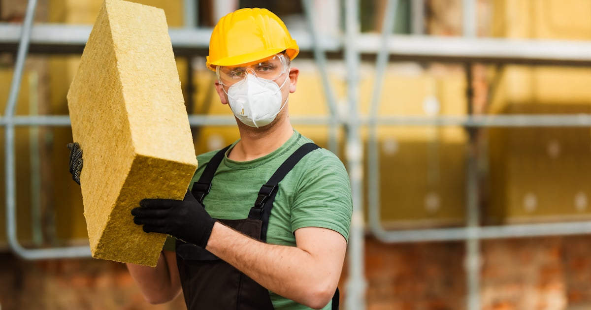 Een isolatiespecialist heeft een stuk steenwol vast. De expert draagt een helm, bril, mondmasker en handschoenen om irritatie van de vezels te voorkomen