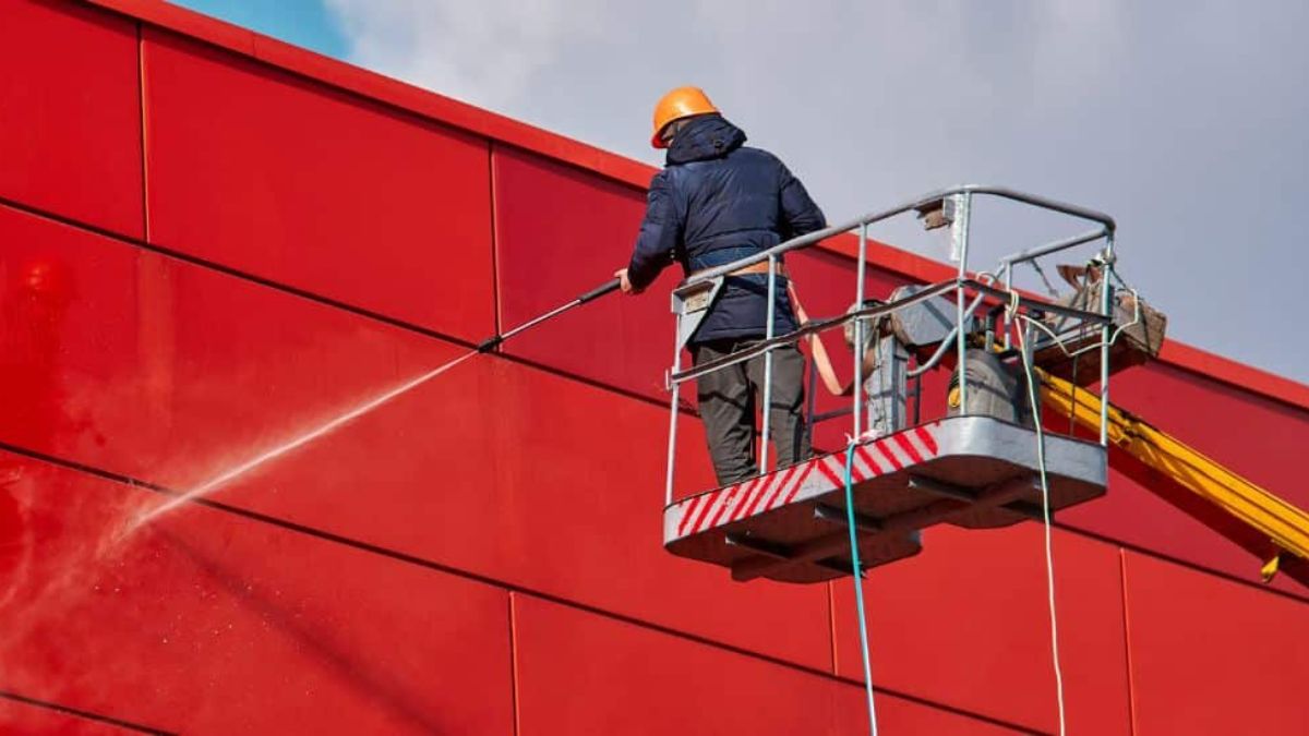 Professional reinigt een rode gevel met een stoom-/hogedrukreiniger vanuit een hoogwerker.