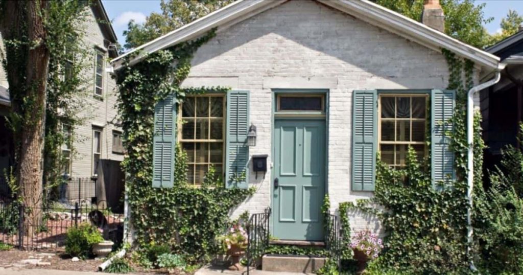 Een klein vrijstaand huis met een blauwe deur en veel begroeiing heeft een witte gevel van sierpleister met een baksteenmotief.