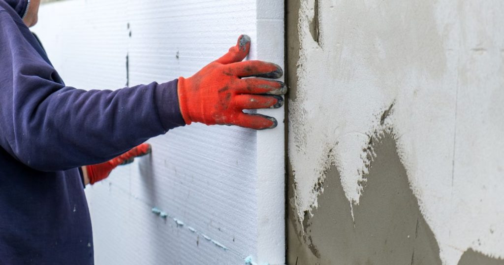 Een vakman plaatst isolatiemateriaal tegen een onafgewerkte gevel. De specialist draagt oranje handschoenen en donkere kleding.